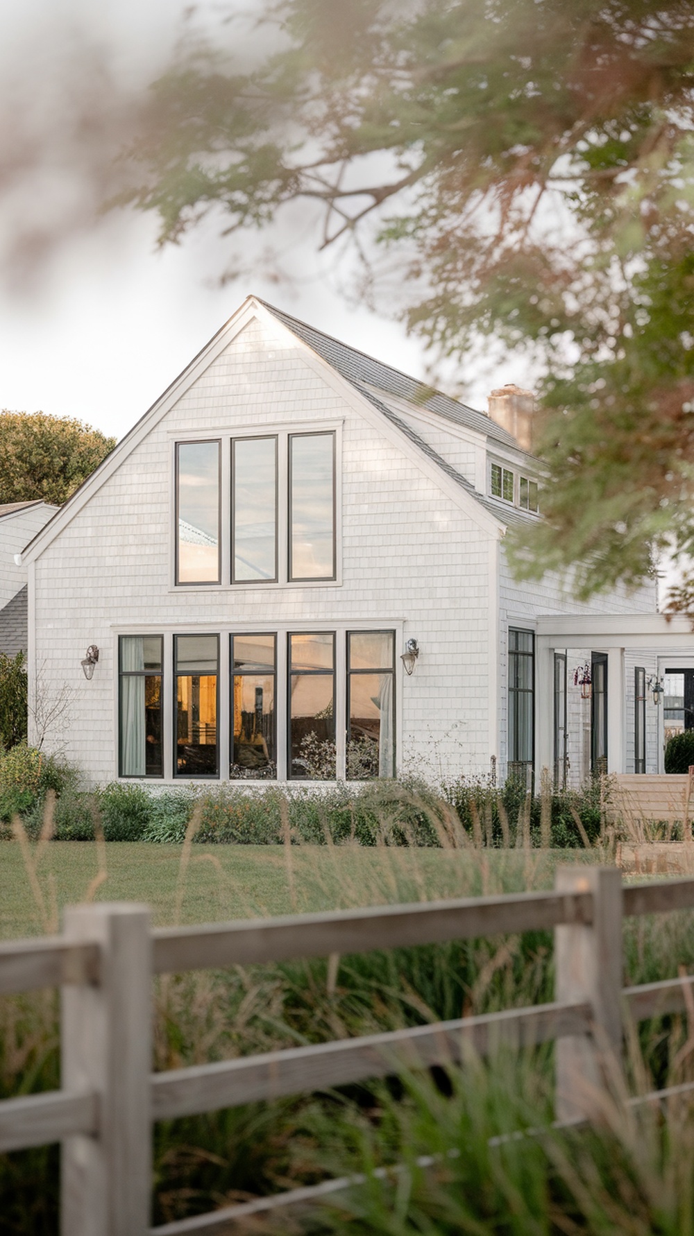 A modern Nantucket style home with white shingles and large windows, surrounded by greenery.