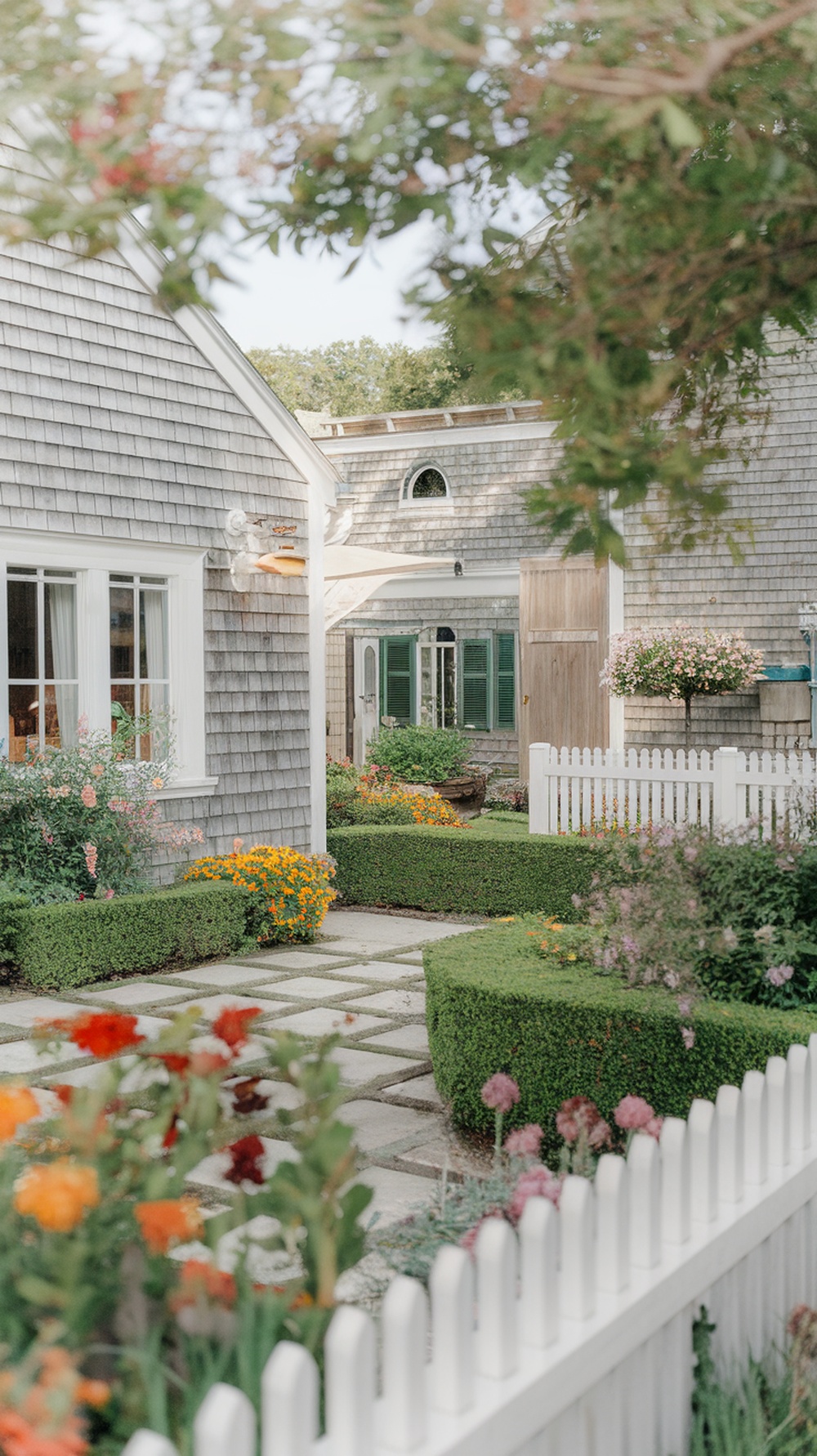 A charming Nantucket style home with lush landscaping, featuring colorful flowers, trimmed hedges, and a stone path.